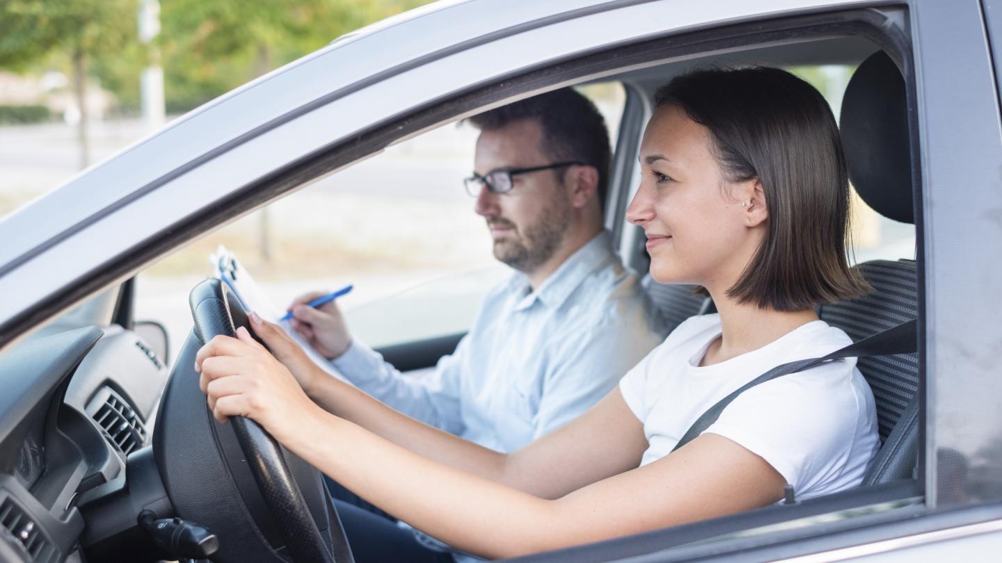 Woman driving a car with a man with a clipboard and pen in the passenger seat