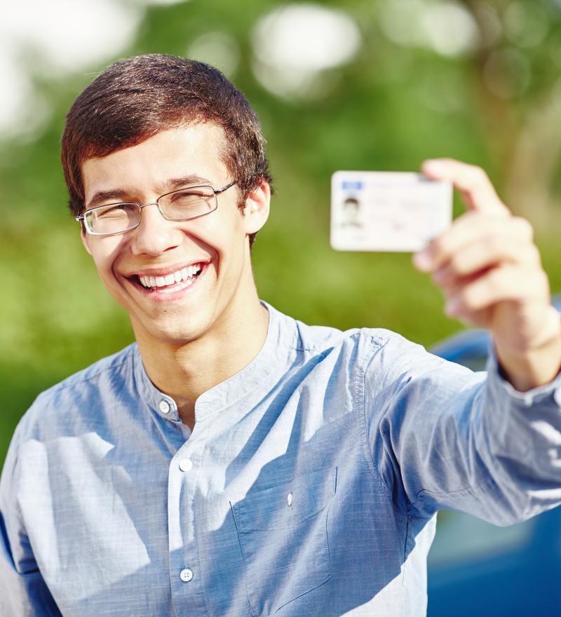 young man in blue shirt and glasses smiling outside holding up driver's license